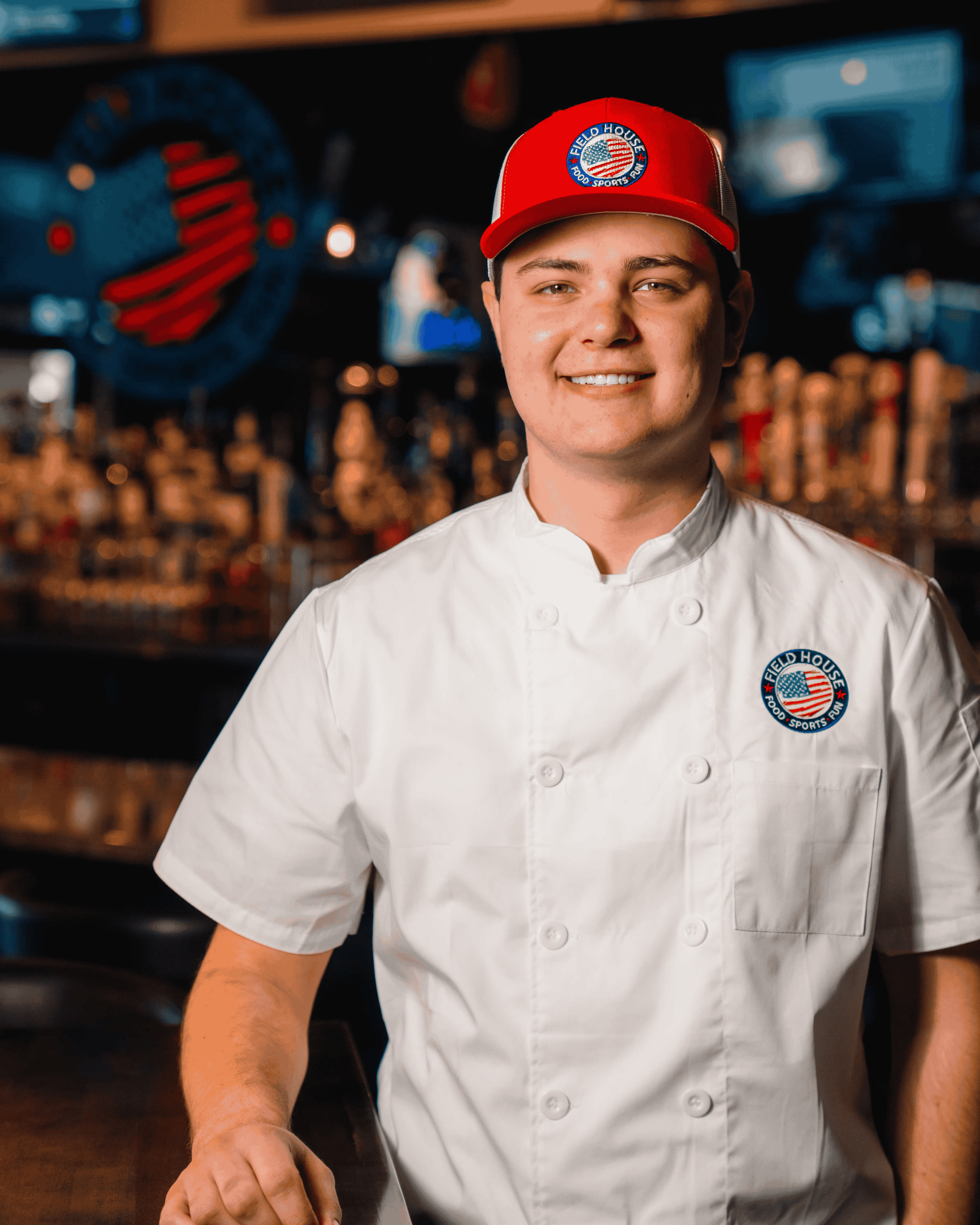 A smiling young man wearing a white chef's coat and a red baseball cap stands in a dimly lit bar. The cap and coat both feature a circular logo with the words Field House Food & Sports Bar and an American flag design. The man has a clean-shaven face and short, brown hair. He is leaning slightly on a counter with his left hand, and his right arm is out of frame. The background is out of focus, showing a well-stocked bar with various bottles of liquor on shelves and several televisions in the distance.