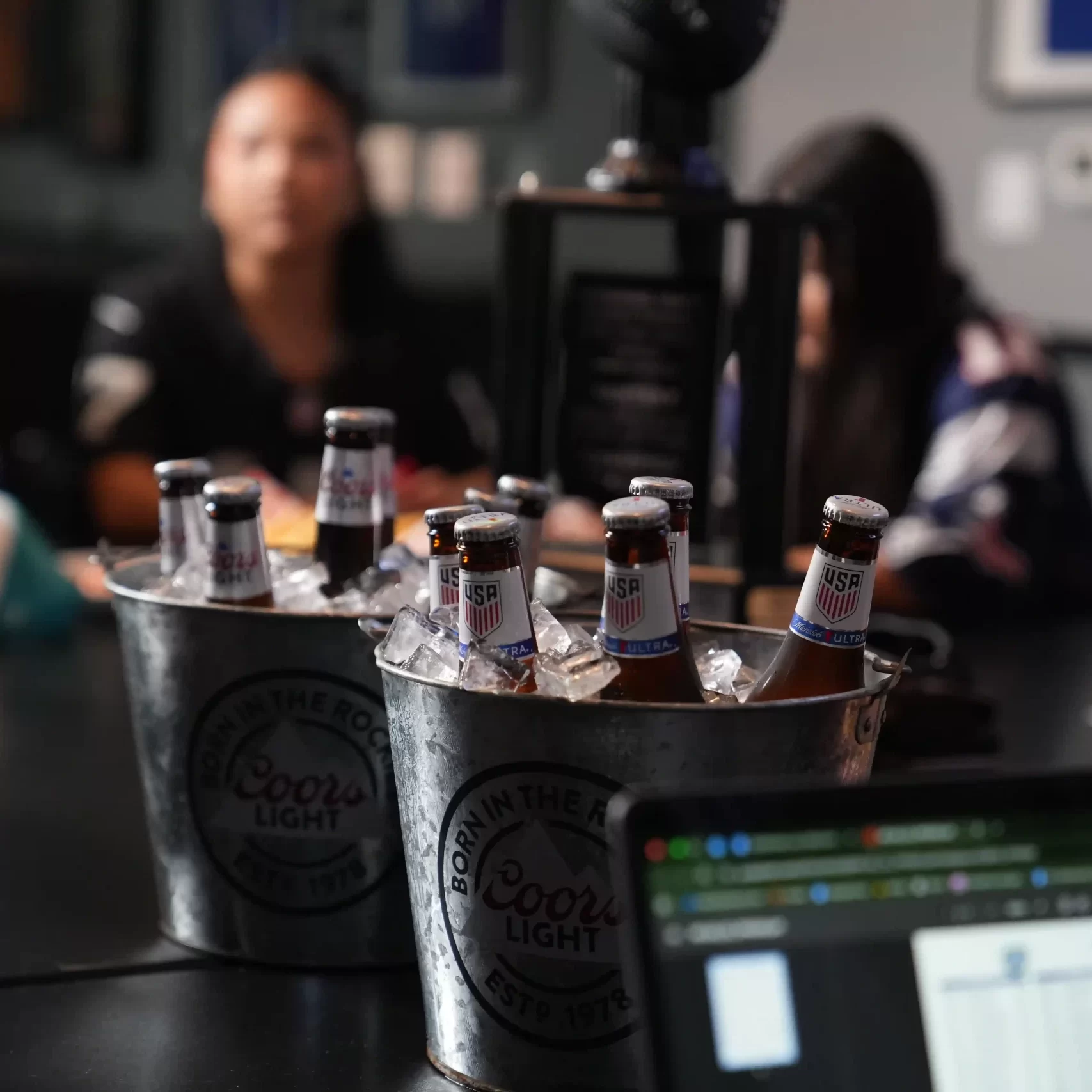 Two buckets of beers with football fans at a large table in the background