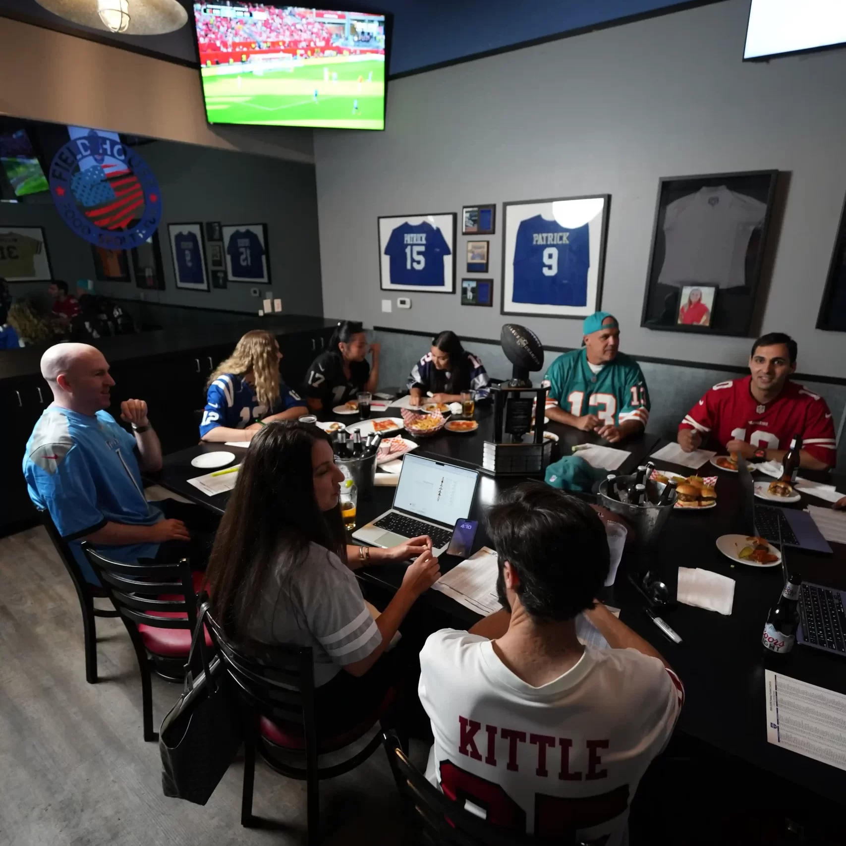 People gathered around a table for a private fantasy football draft party with appetizers, trophy, beers, laptops having a good time